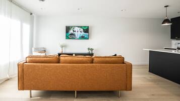 Een moderne woonkamer met een bruine leren bank, een klein bijzettafeltje met een plant, en een keukenhoek met een zwart aanrecht en hanglampen.