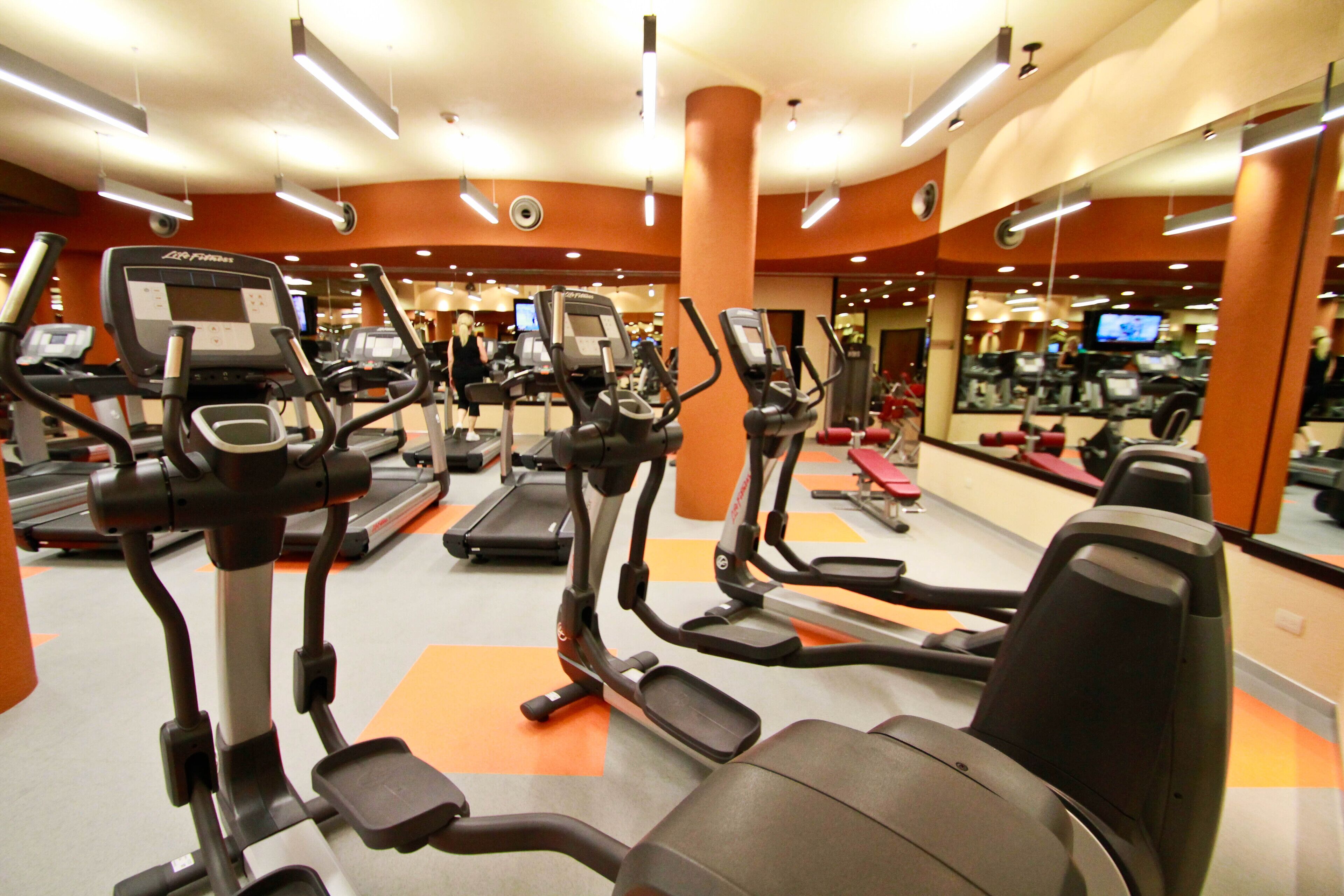 A gym with various exercise machines, including treadmills and elliptical trainers, arranged in a spacious layout with orange flooring and mirrored walls.