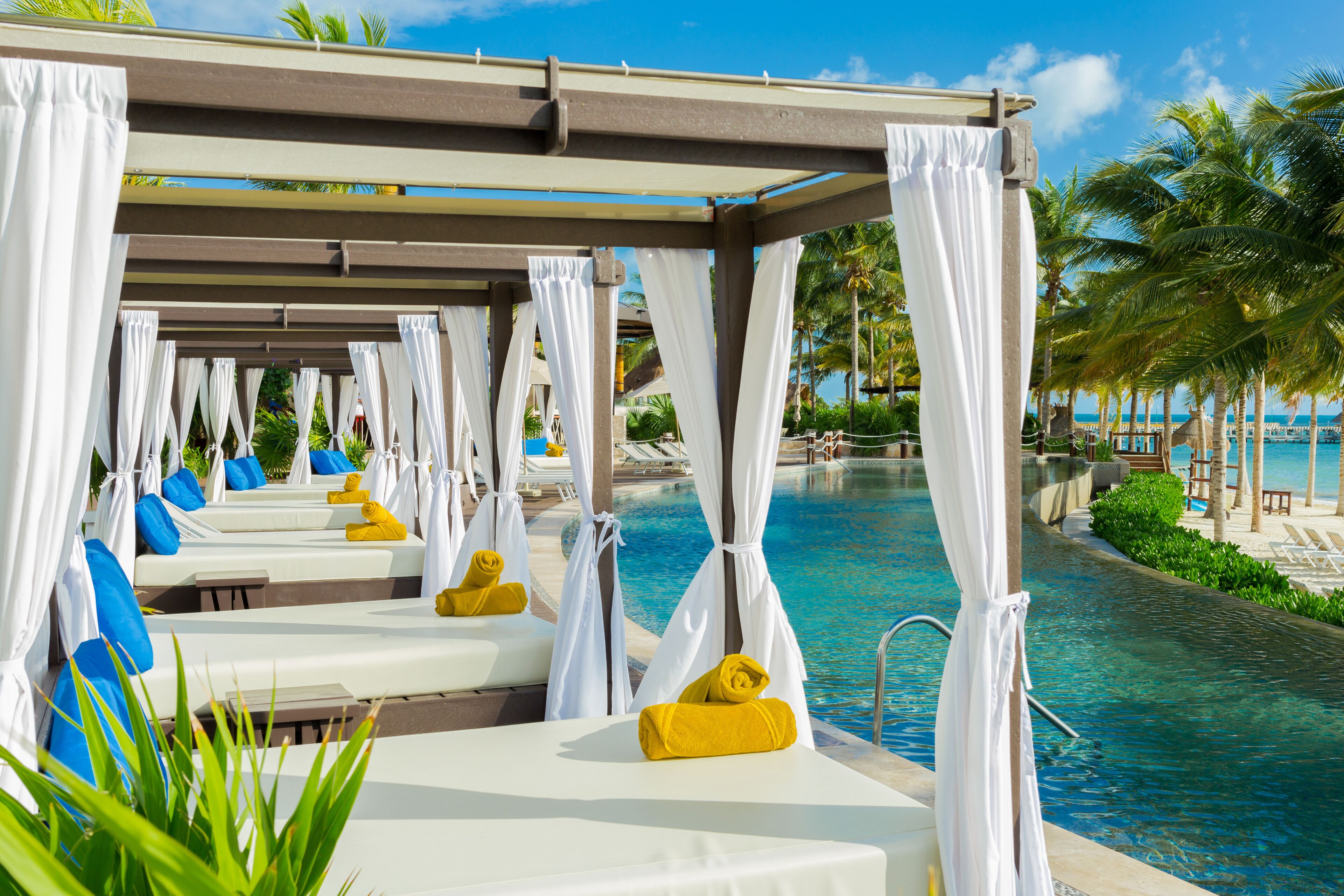 A poolside area with white canopies, yellow towels, and blue cushions on lounge chairs.