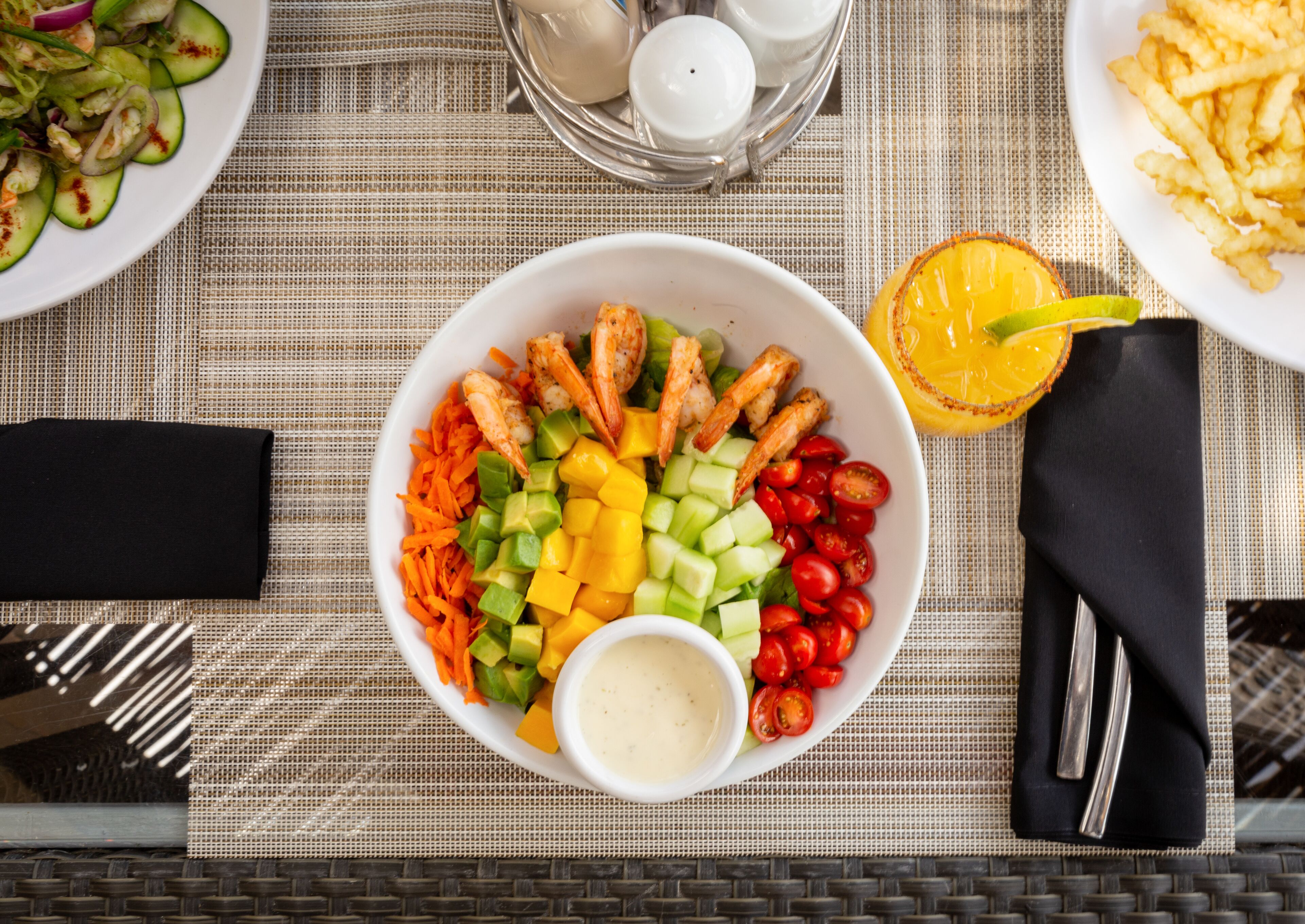 Una mesa con un bol de verduras y camarones, un vaso de jugo de naranja y un plato de papas fritas.
