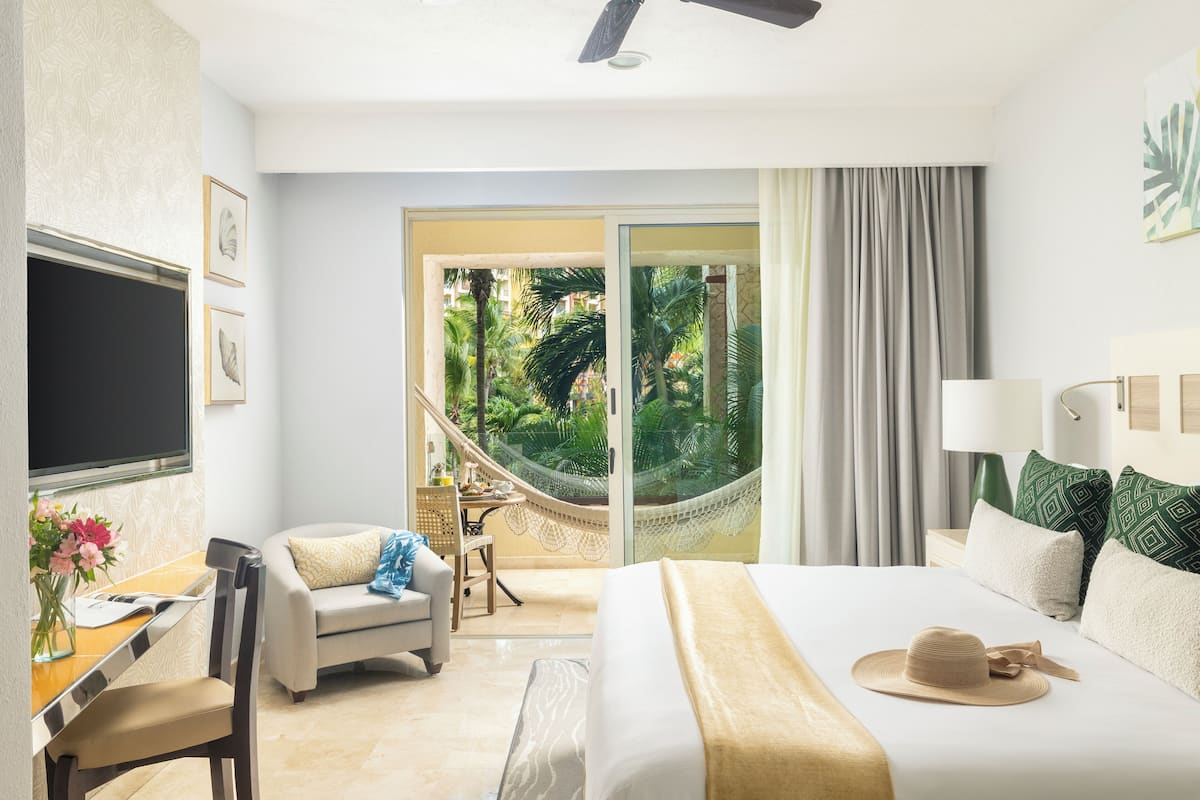 Habitación de hotel con cama, escritorio, silla, televisor y vista a un jardín a través de un ventanal.