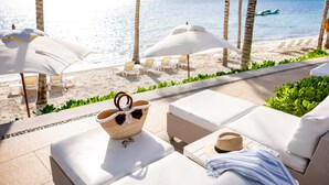 Un área de descanso en la playa con asientos blancos acolchados, una cesta de mimbre y un sombrero de paja sobre una mesa.