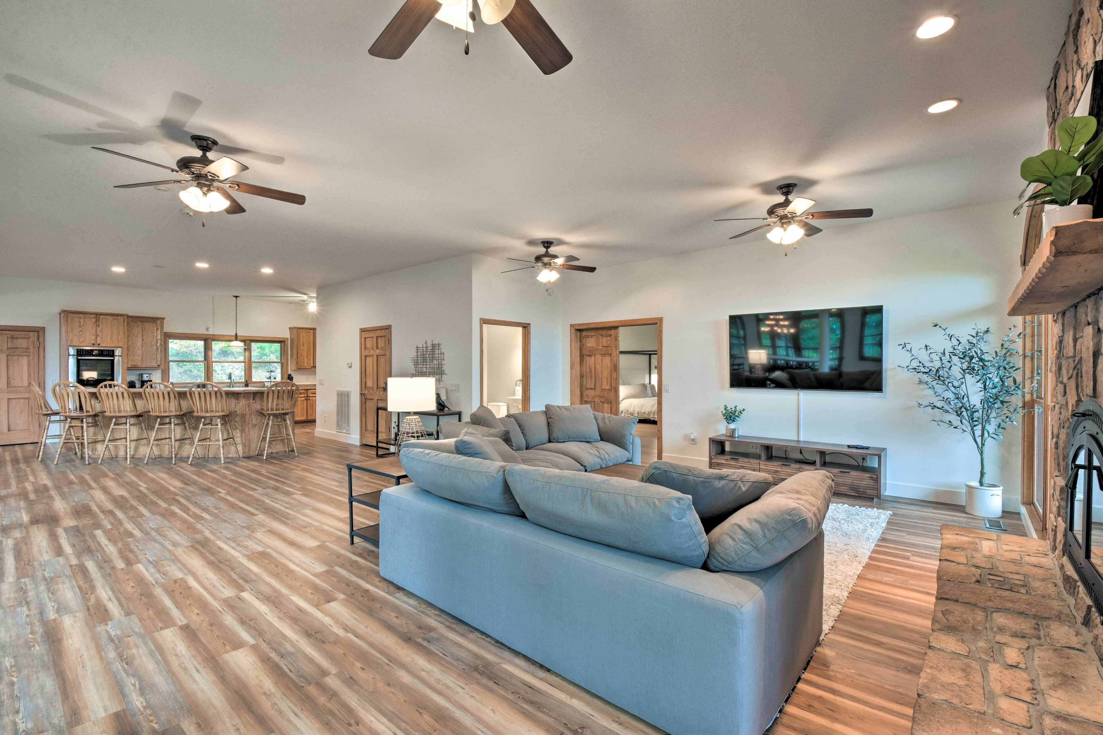 A spacious living area with a large sofa, wooden flooring, ceiling fans, and a kitchen in the background.