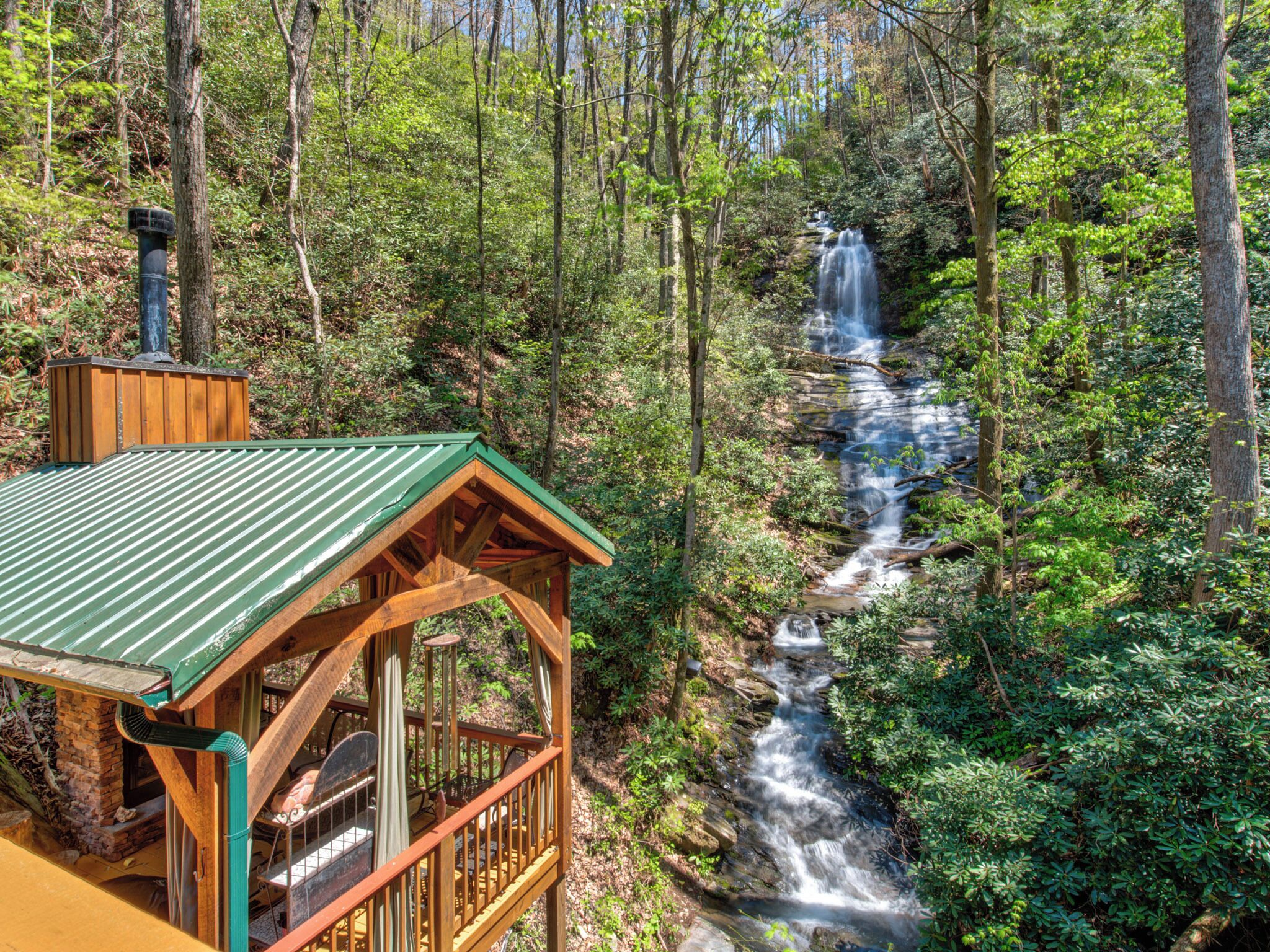 A wooden cabin with a green roof overlooking a waterfall in a forest.
