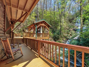 Um deck de madeira com uma cadeira de balanço, uma pequena cabana e vista para uma área florestal com uma cascata.