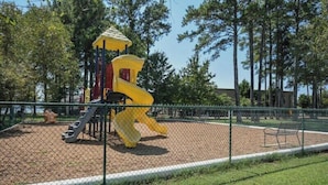 A playground with a yellow slide and climbing structures, surrounded by a fence and trees. - Dockside at River Rocks Landing (Gadsden)