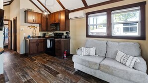 A kitchen with wooden cabinets, a microwave, and a stove. There is a gray sofa with patterned cushions and a window with a view of the outside. - Dockside at River Rocks Landing (Gadsden)