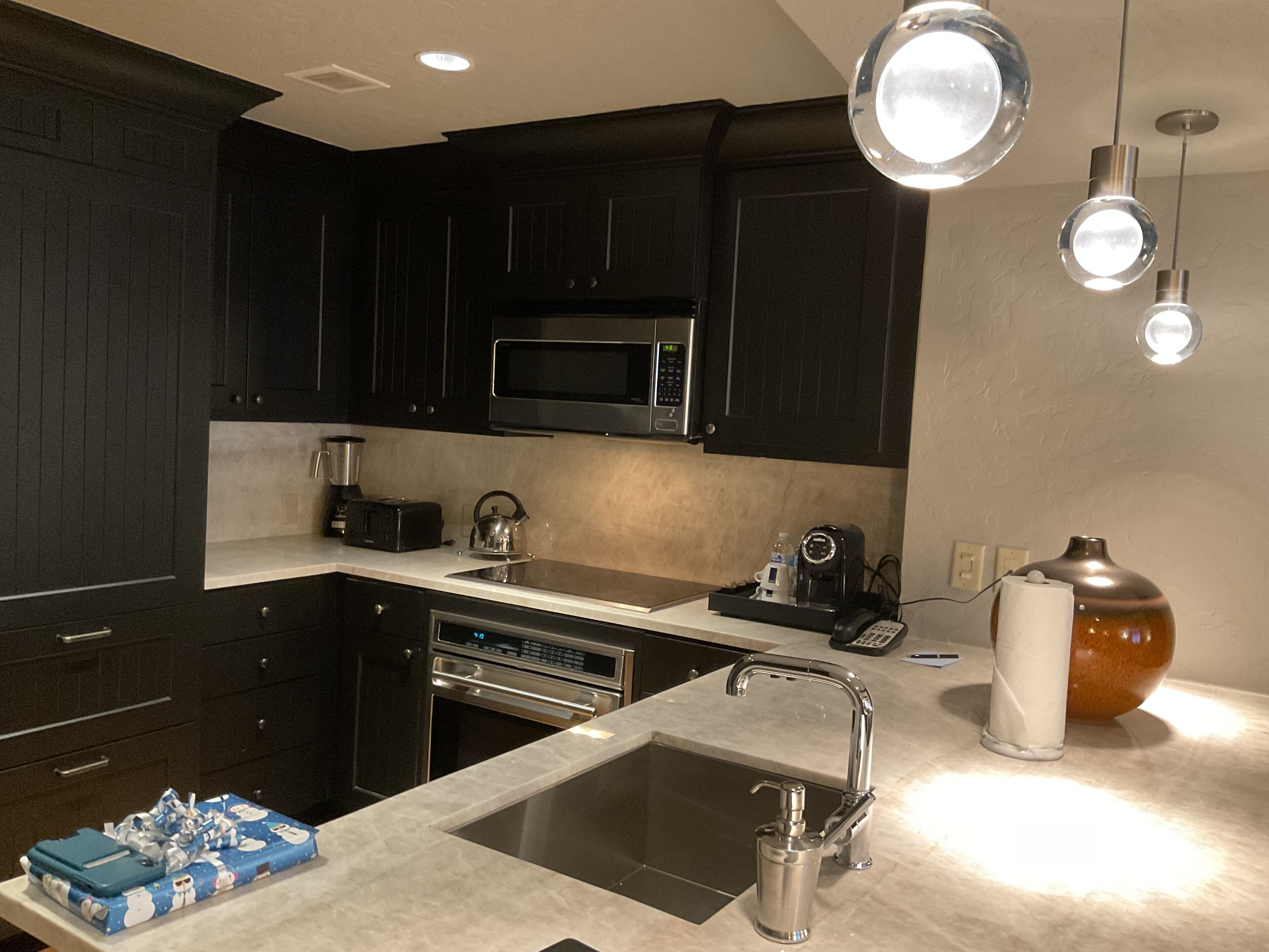 Una cocina moderna con armarios oscuros, un microondas de acero inoxidable, una cafetera y un lavabo.