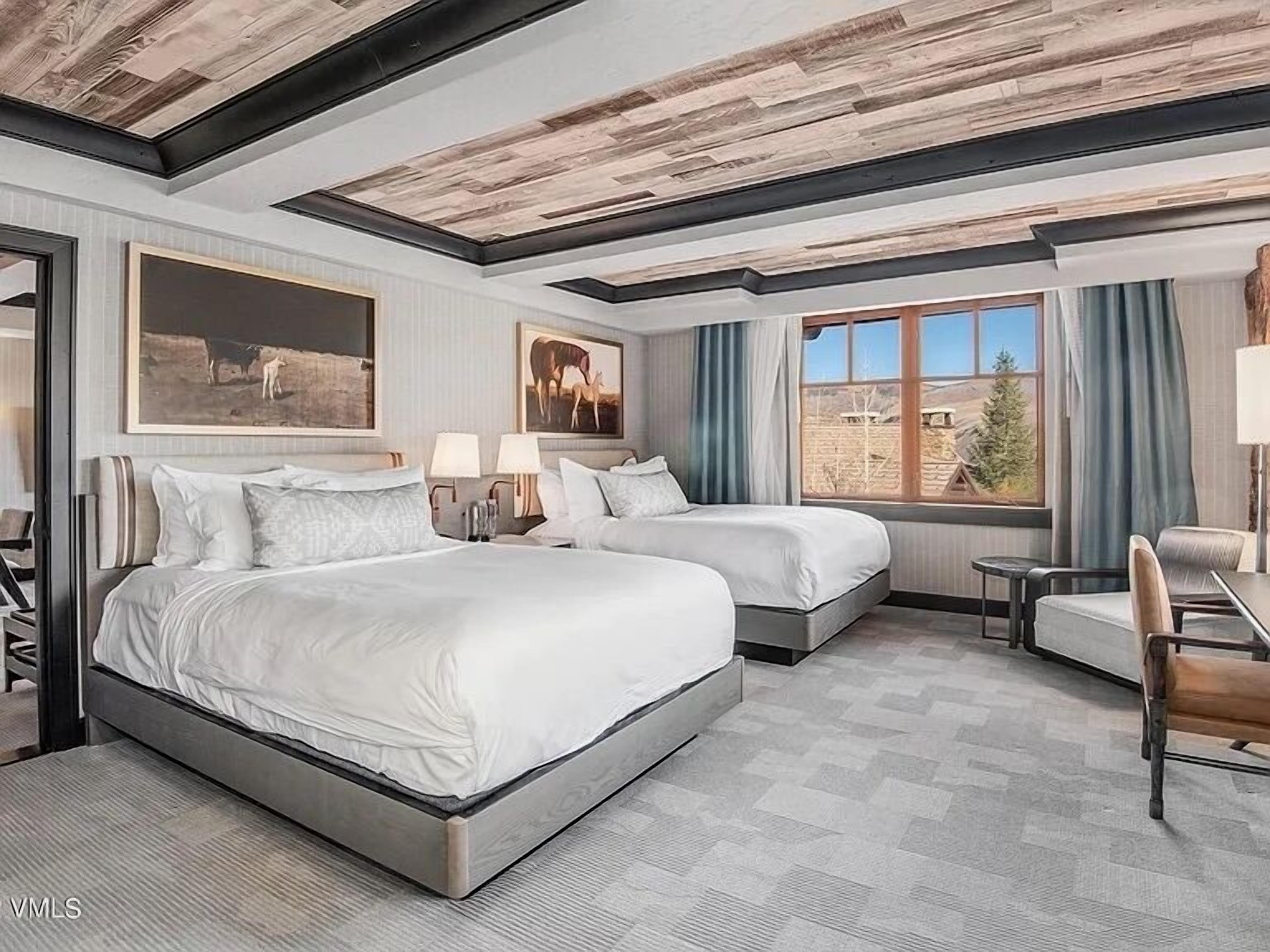 Habitación de hotel con dos camas, un escritorio y una silla. Hay dos cuadros en la pared y una ventana con cortinas.