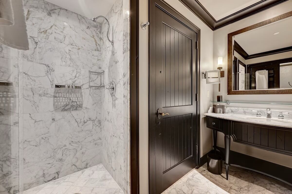 Un baño moderno con pared de ducha de mármol, puerta de madera oscura y un espejo grande sobre un tocador con encimera de cristal.