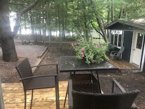 A wooden deck with two wicker chairs and a table, surrounded by a garden with a small shed and a tree. - Lake Rosseau peace and Serenity  (Rosseau)
