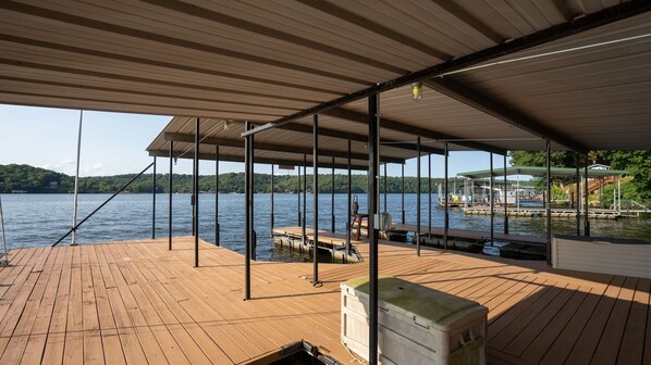 A wooden deck with a covered area, a dock extending into the water, and a view of a lake with hills in the background.
