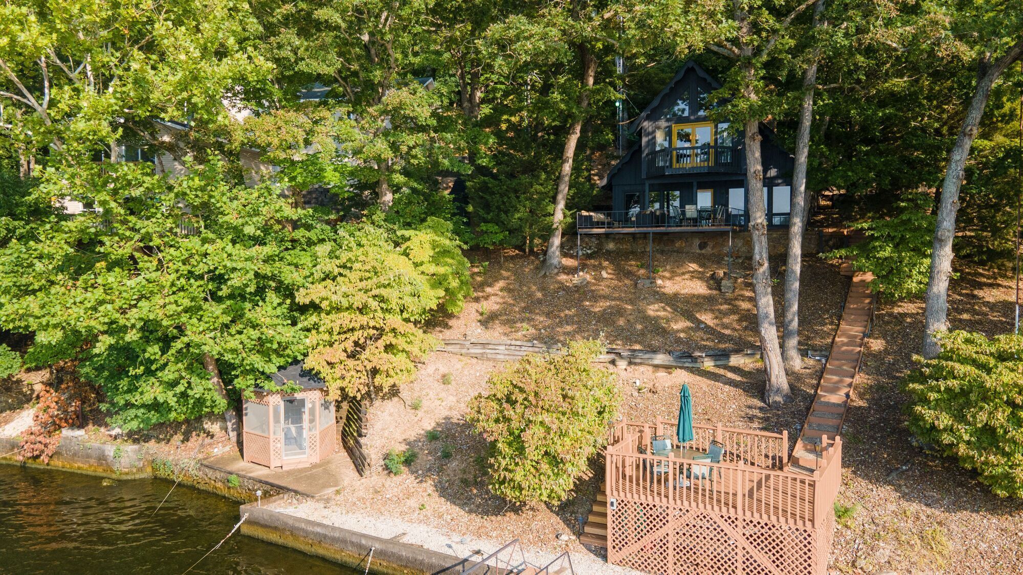 A cabin with a deck, surrounded by dense trees and a body of water.