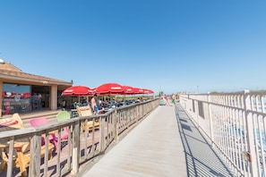 Une promenade en bois avec des espaces de repos en plein air et des parasols rouges, qui mène à une plage.