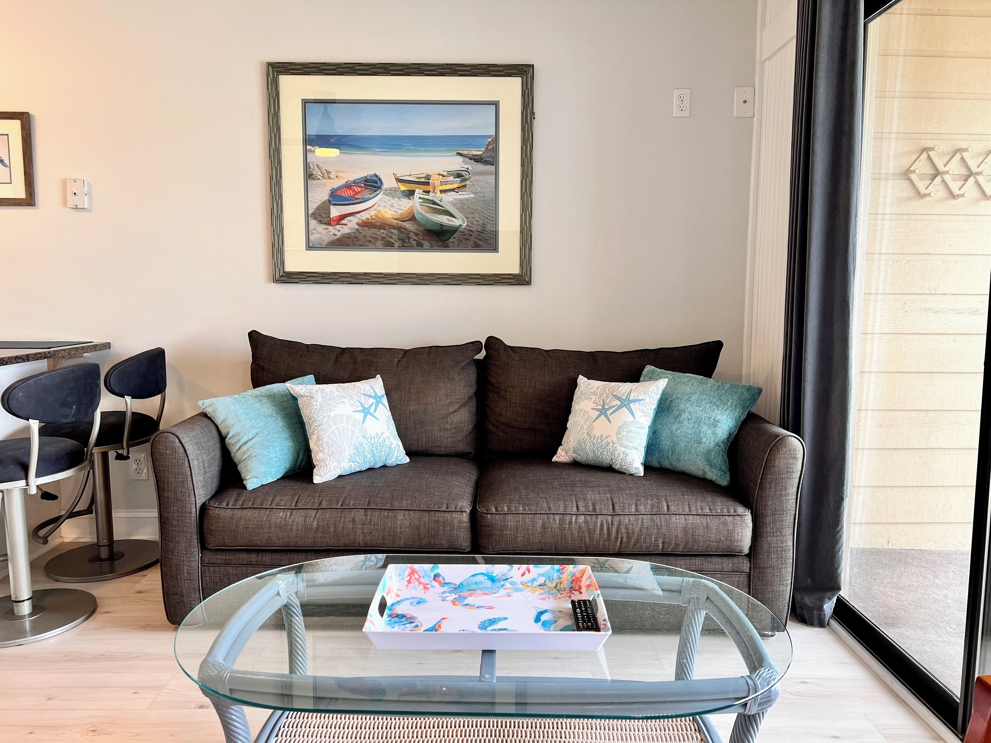 A living room with a brown sofa, two blue and white patterned pillows, a glass coffee table with a colorful tray, and a framed painting of boats on a beach.