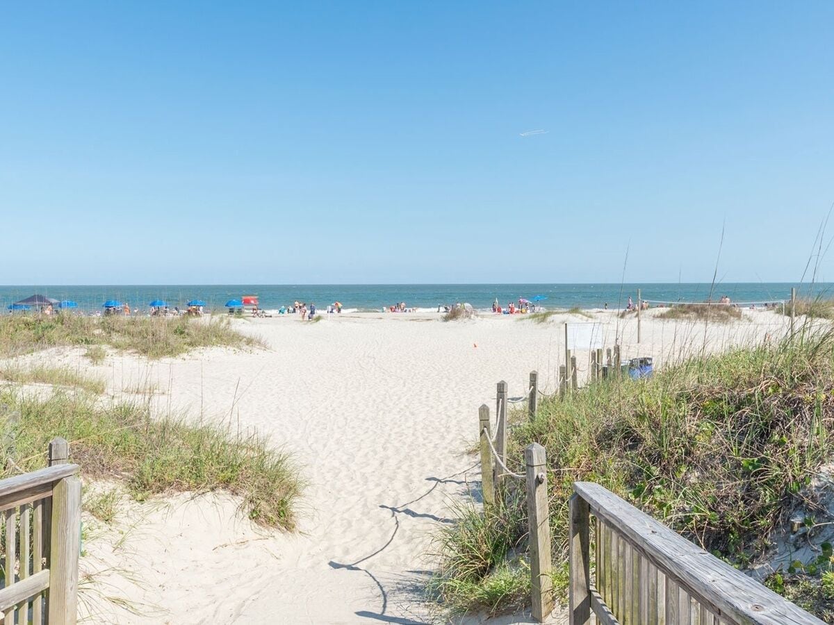 Une plage de sable avec une promenade en bois menant à l’eau, et des parasols de plage visibles au loin.