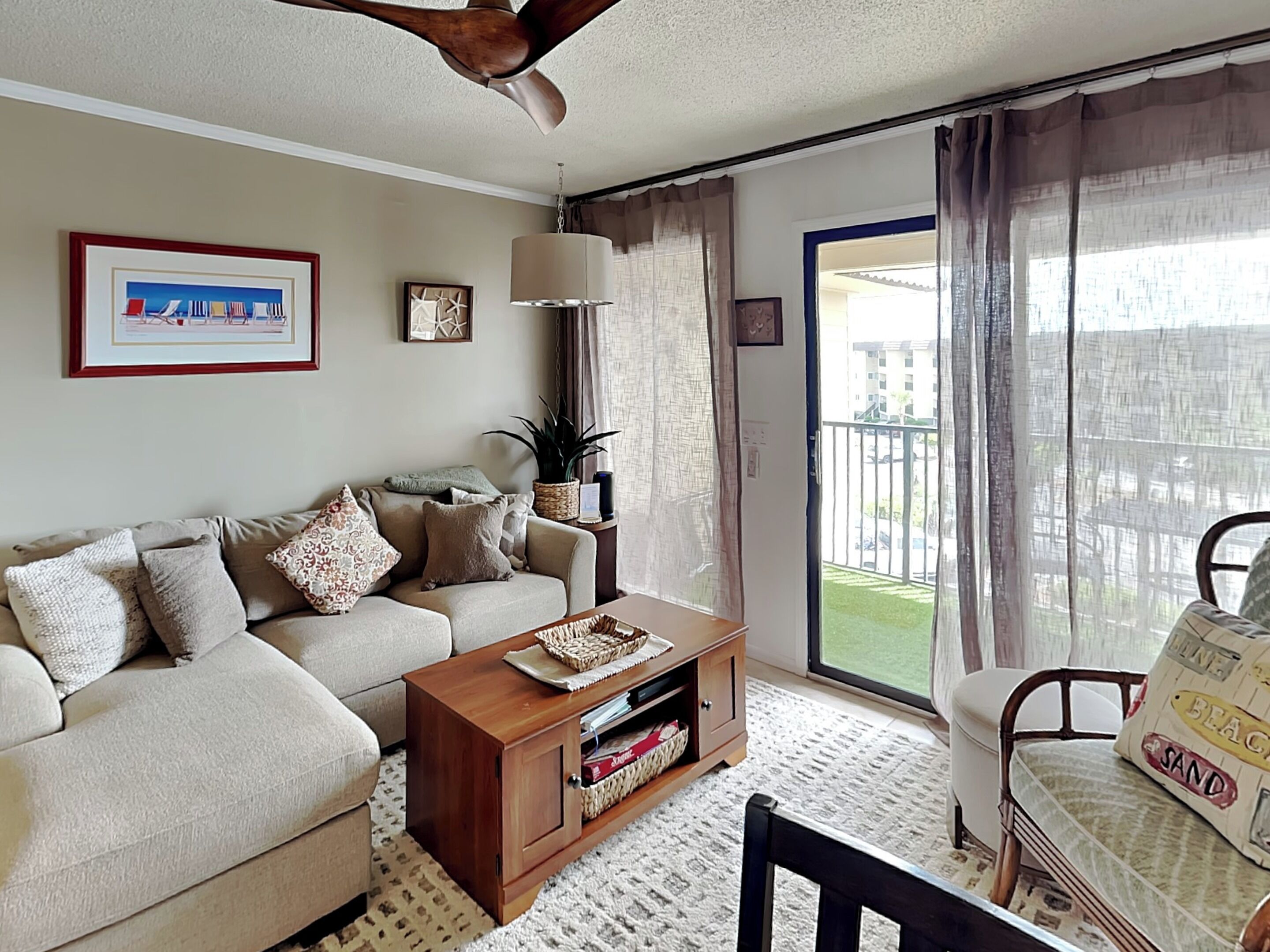 A living room with a sofa, coffee table, and a chair. There is a ceiling fan, a window with curtains, and a view of a building outside.