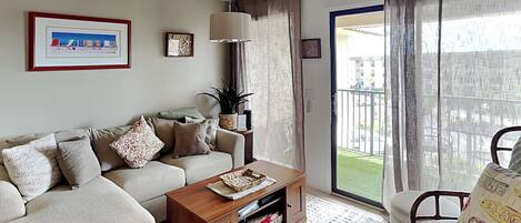 A living room with a sofa, coffee table, and a chair. There is a ceiling fan, a window with curtains, and a view of a building outside.