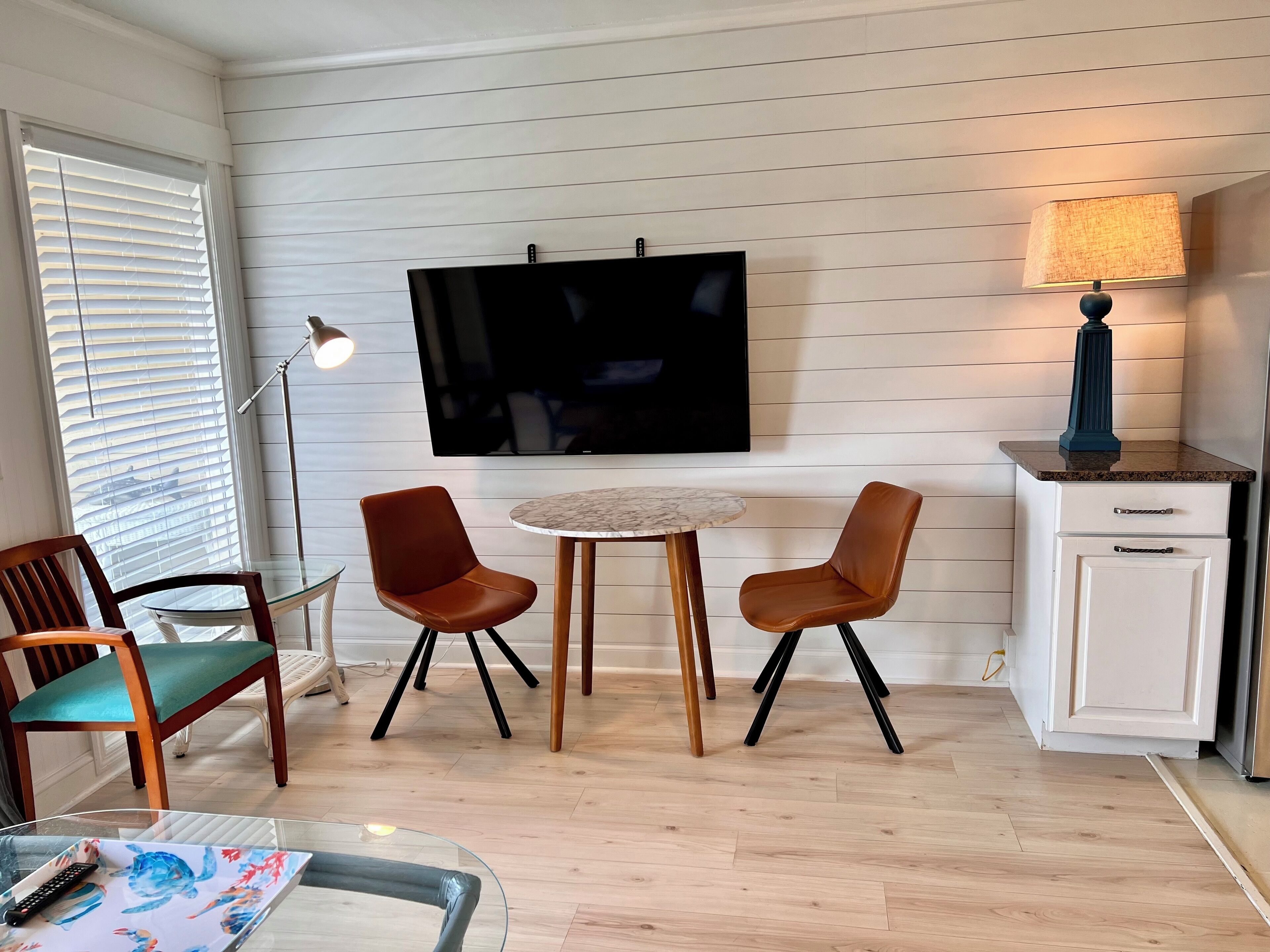 Ett modernt vardagsrum med en platt-TV, två bruna läderfåtöljer, ett soffbord i glas och ett litet vitt skåp med en lampa.