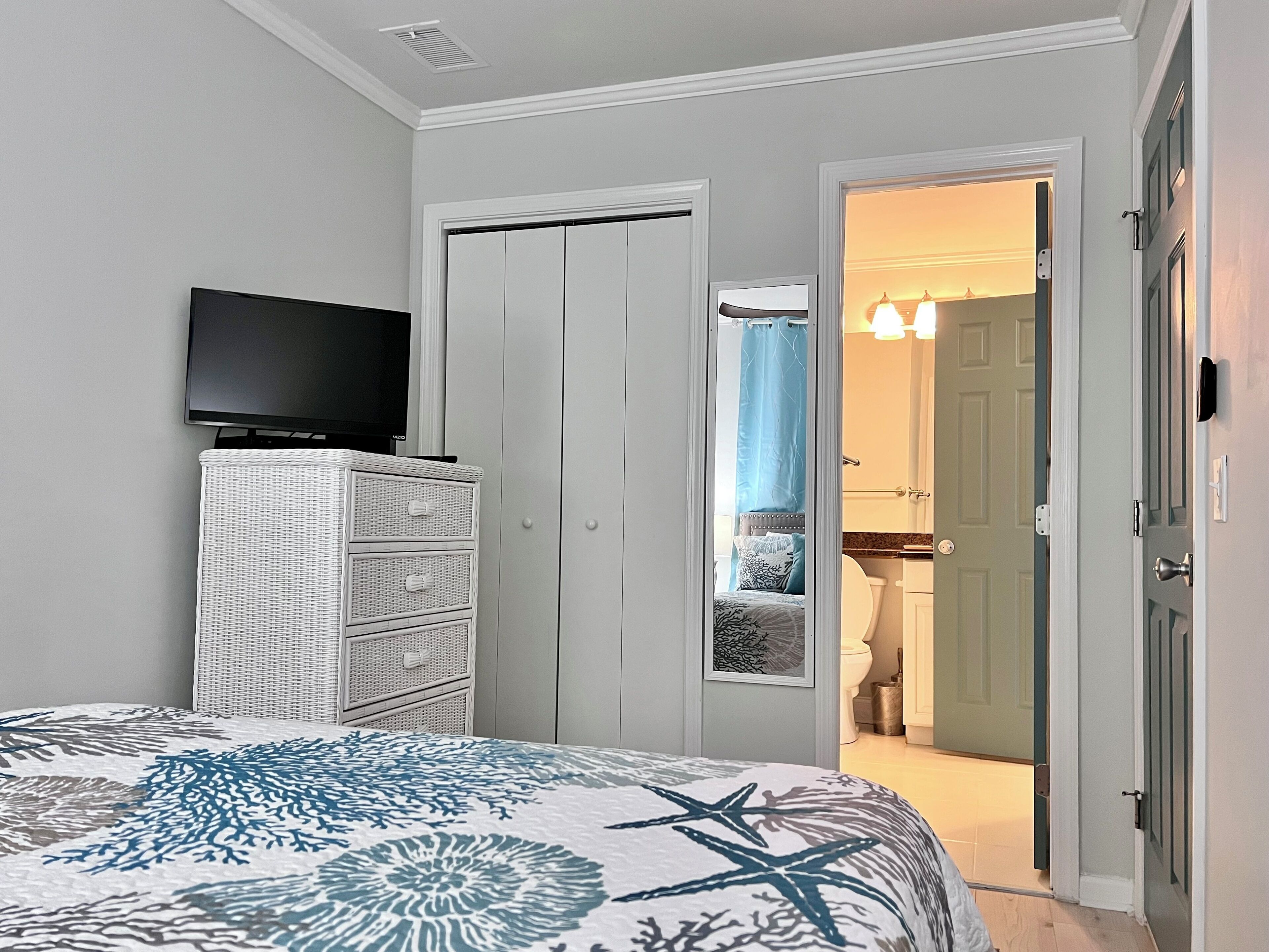 Une chambre à coucher comprenant un lit, un téléviseur, une commode en osier blanche, une porte donnant sur une salle de bain et un couloir.
