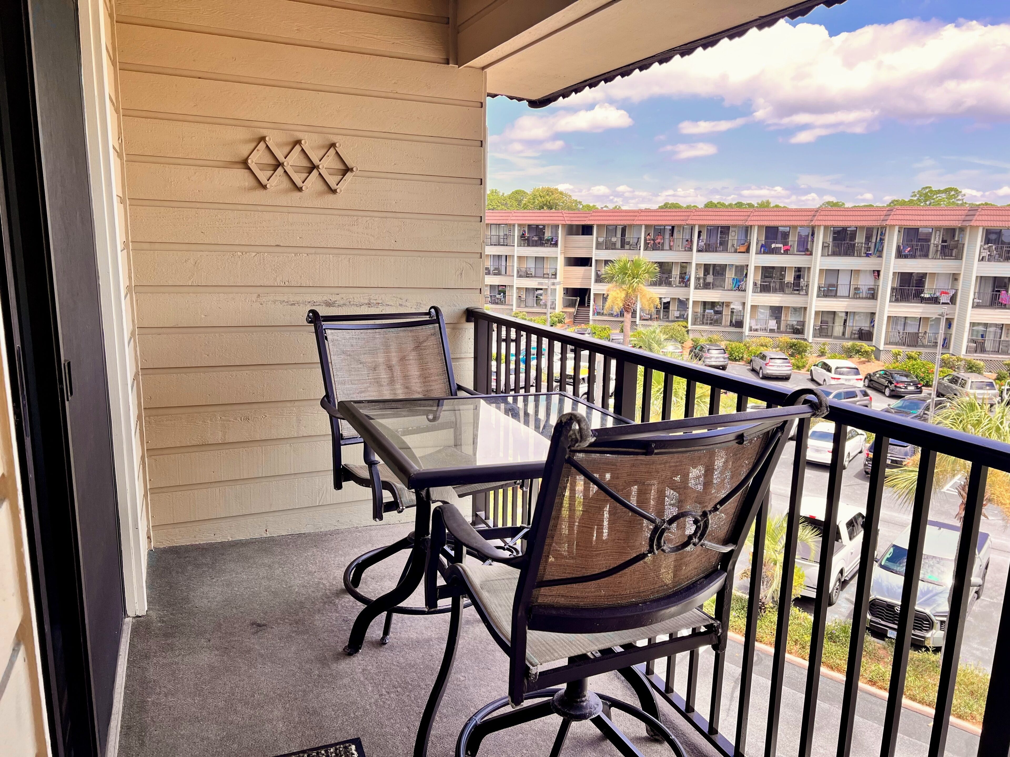 A balcony with a glass table and two chairs, overlooking a parking area and a building with multiple balconies.