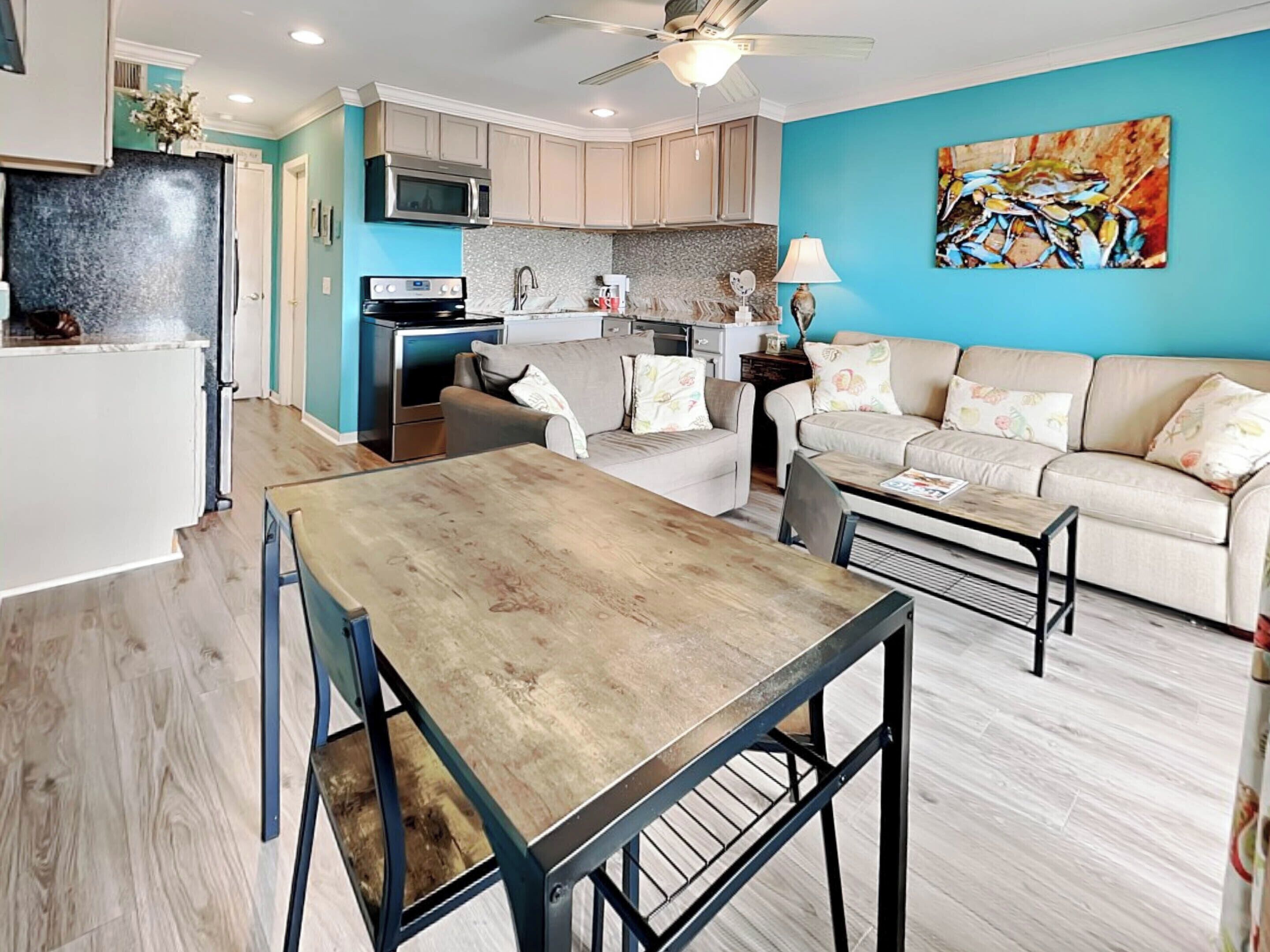 A modern kitchen with a microwave, oven, and sink. There is a dining area with a wooden table and chairs. A sofa and a coffee table are in the living area.