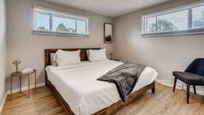 Een slaapkamer met een houten bed, witte beddengoed, twee ramen met zonwering, een nachtkastje met een vaas, en een stoel.