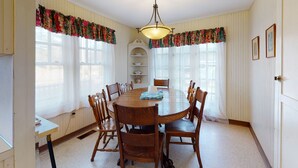 Uma sala de jantar com uma mesa redonda de madeira, rodeada por cadeiras, um lustre e cortinas com detalhes florais.