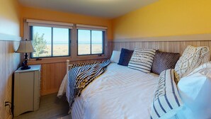 Quarto com cama, mesa de cabeceira, abajur e janela com vista para a praia.