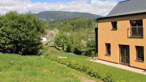 A two-story house with a balcony, surrounded by lush greenery and a mountainous backdrop.
