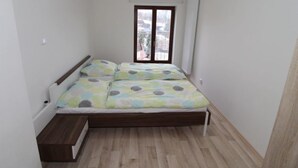 A small, single-bed room with a wooden floor, a window with a view of buildings, and a white wall.