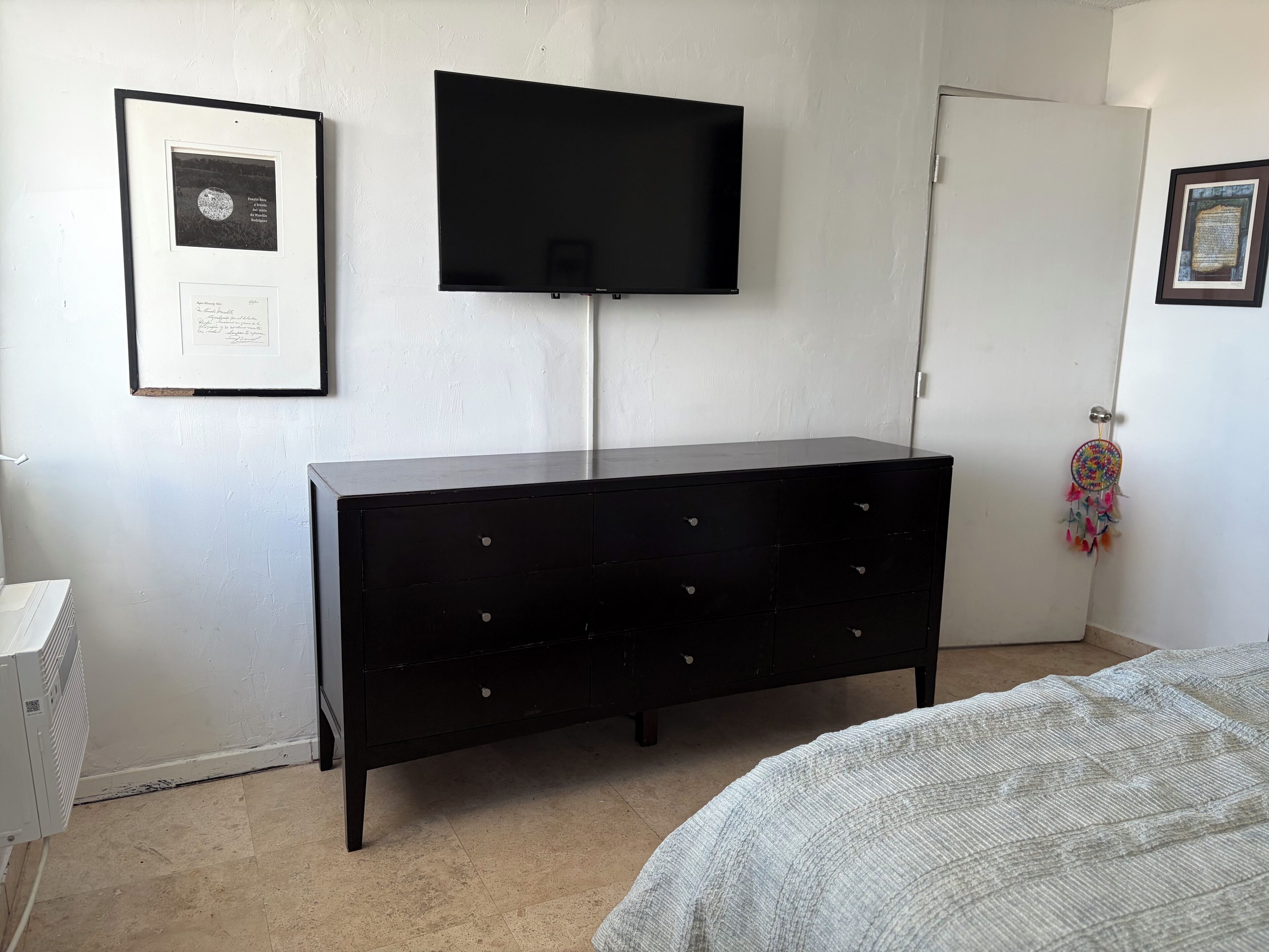 Une chambre avec une commode noire, un téléviseur à écran plat fixé au mur et une porte décorée d’un attrape-rêves.