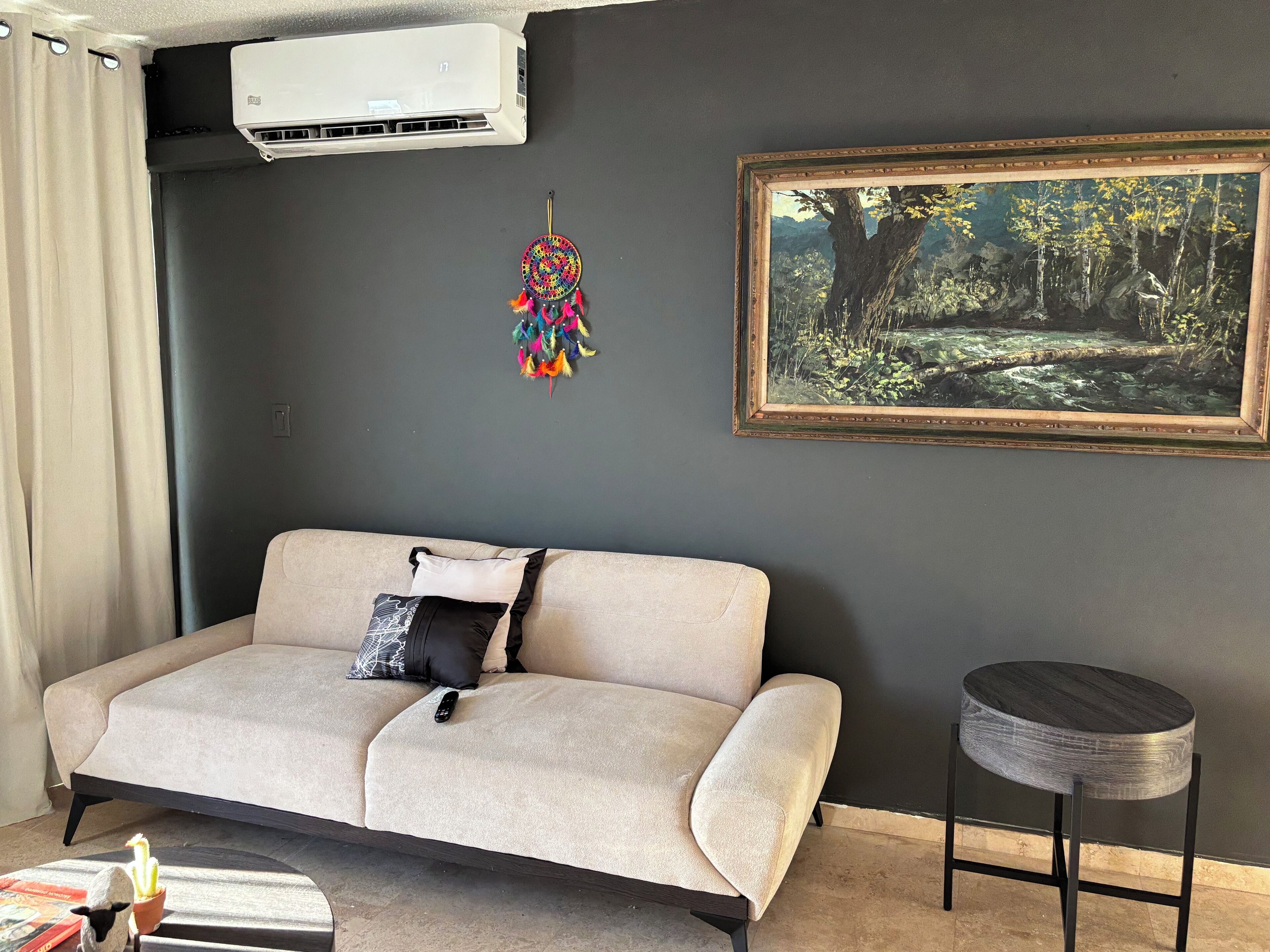 Ein Wohnzimmer mit einer Couch, einem kleinen runden Tisch, einer Wand-Klimaanlage, einem gerahmten Gemälde und einem Traumfänger.