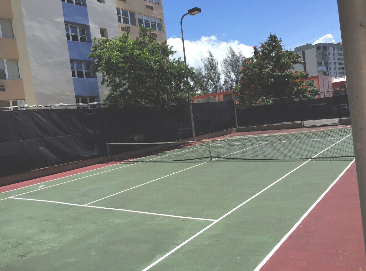 An outdoor tennis court with a net, surrounded by a fence, and buildings in the background.