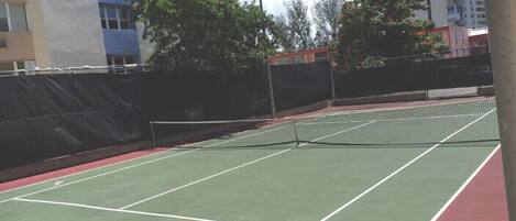An outdoor tennis court with a net, surrounded by a fence, and buildings in the background.