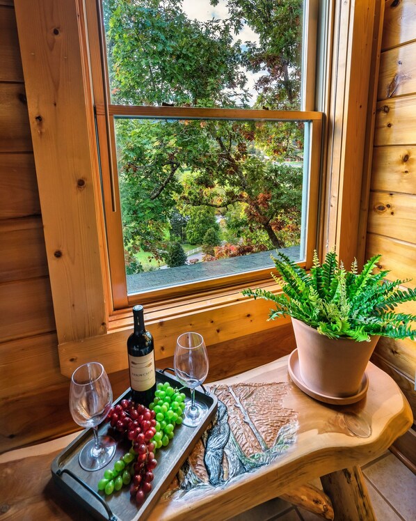 A wooden table with a wine bottle, two glasses, and a tray of grapes by a window with a view of trees.