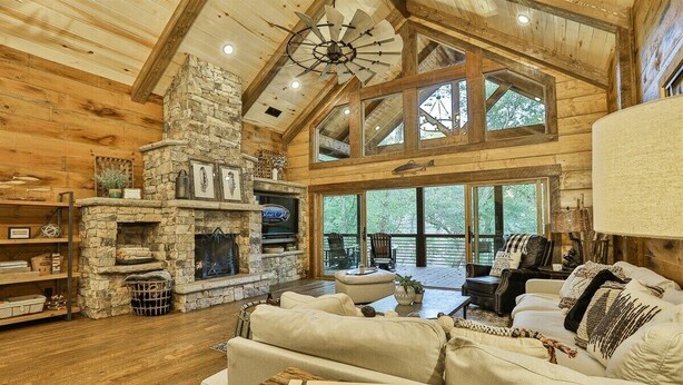 A rustic cabin interior with a stone fireplace, wooden beams, a large window, and a guest book on a table. - Riverscape | Ellijay, GA (Ellijay)