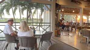 Un espacio de comedor al aire libre con sillas y mesas de mimbre, amplias ventanas que ofrecen vistas a palmeras y un escenario para música en vivo.
