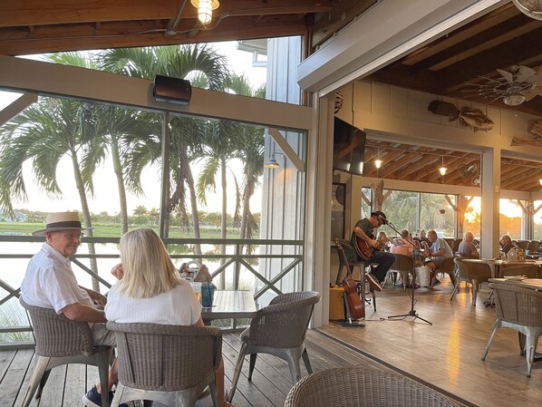 Een buitenruimte met een eethoek, rieten stoelen en tafels, grote ramen met uitzicht op palmbomen en een podium voor live muziek.
