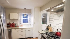 A compact kitchen with white cabinets, a stainless steel refrigerator, a microwave, a gas stove, and a window with patterned curtains.