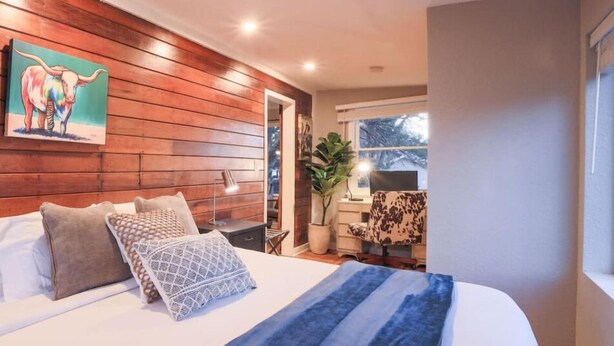 A bedroom with a wooden wall, a bed with pillows, a painting of a bull, and a window with a plant outside.