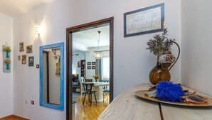 A room with a wooden floor, a mirror with a blue frame, a vase with flowers, and a dining area visible through an open door.