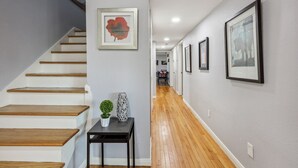 A hallway with a wooden staircase, a small table with a plant, and framed artwork on the walls. - 5BR w/ Gameroom & 1K WiFi - Med Ctr, Downtown, NRG - Sleeps 15 (Houston)