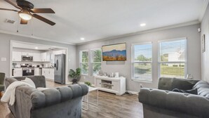 A modern living room with grey sofas, a white entertainment unit, and a kitchen in the background. - 5BR w/ Gameroom & 1K WiFi - Med Ctr, Downtown, NRG - Sleeps 15 (Houston)