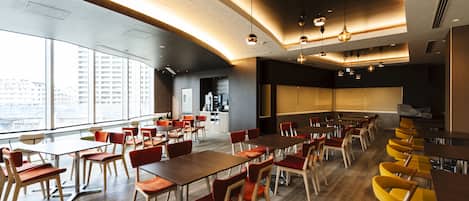 A modern dining area with multiple tables and chairs, large windows, and a kitchen in the background.
