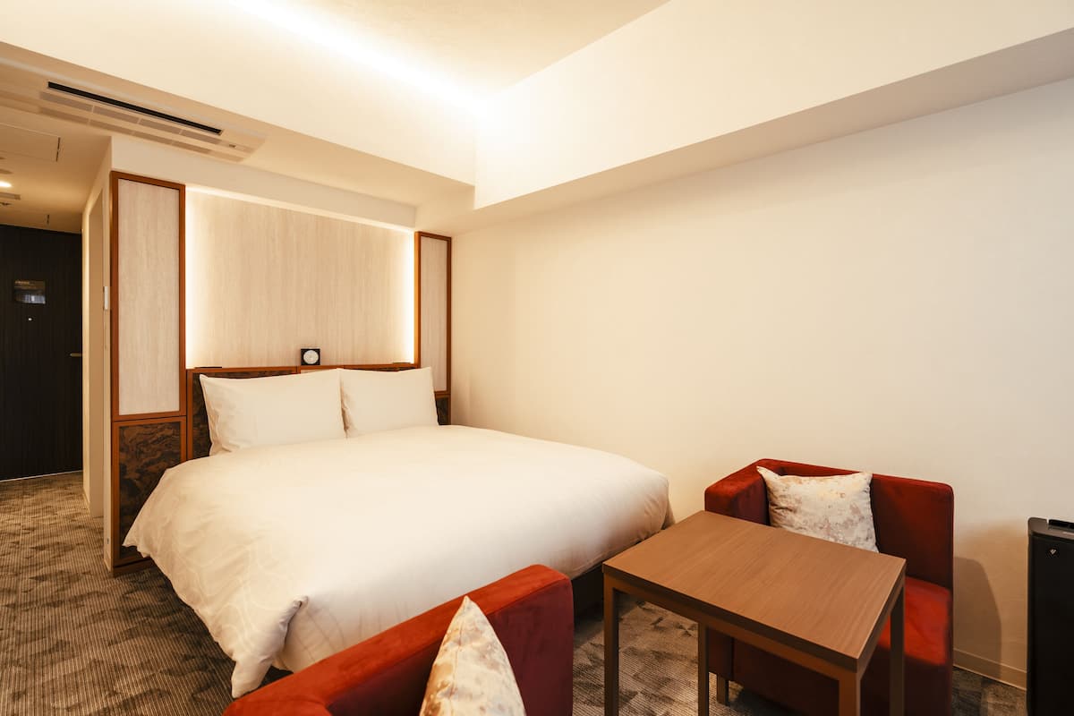A hotel room with a large bed, two red armchairs, a small wooden table, and a wall-mounted TV.