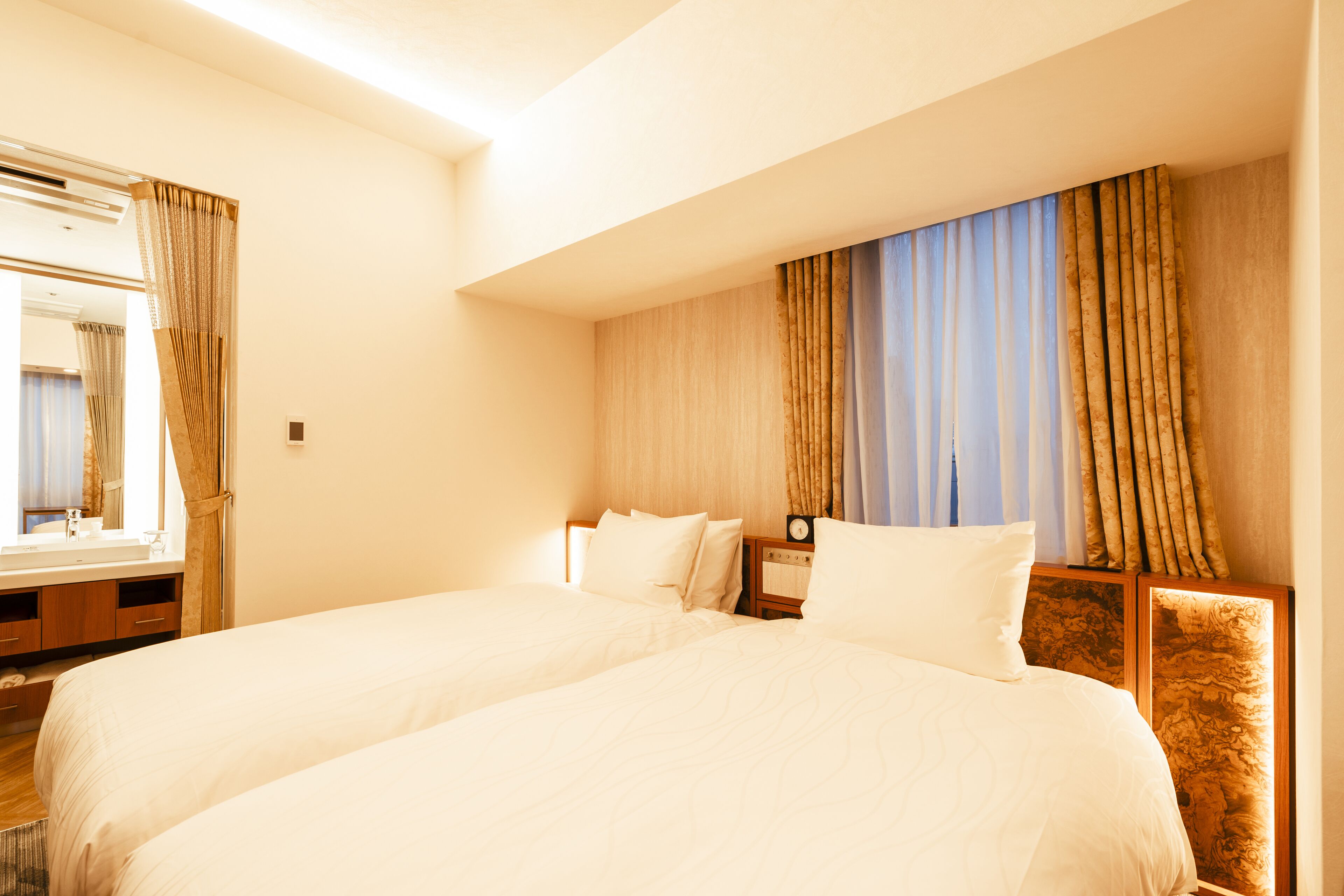 Quarto de hotel com duas camas, cabeceira de madeira, mesa de cabeceira com relógio e janela com cortinas.
