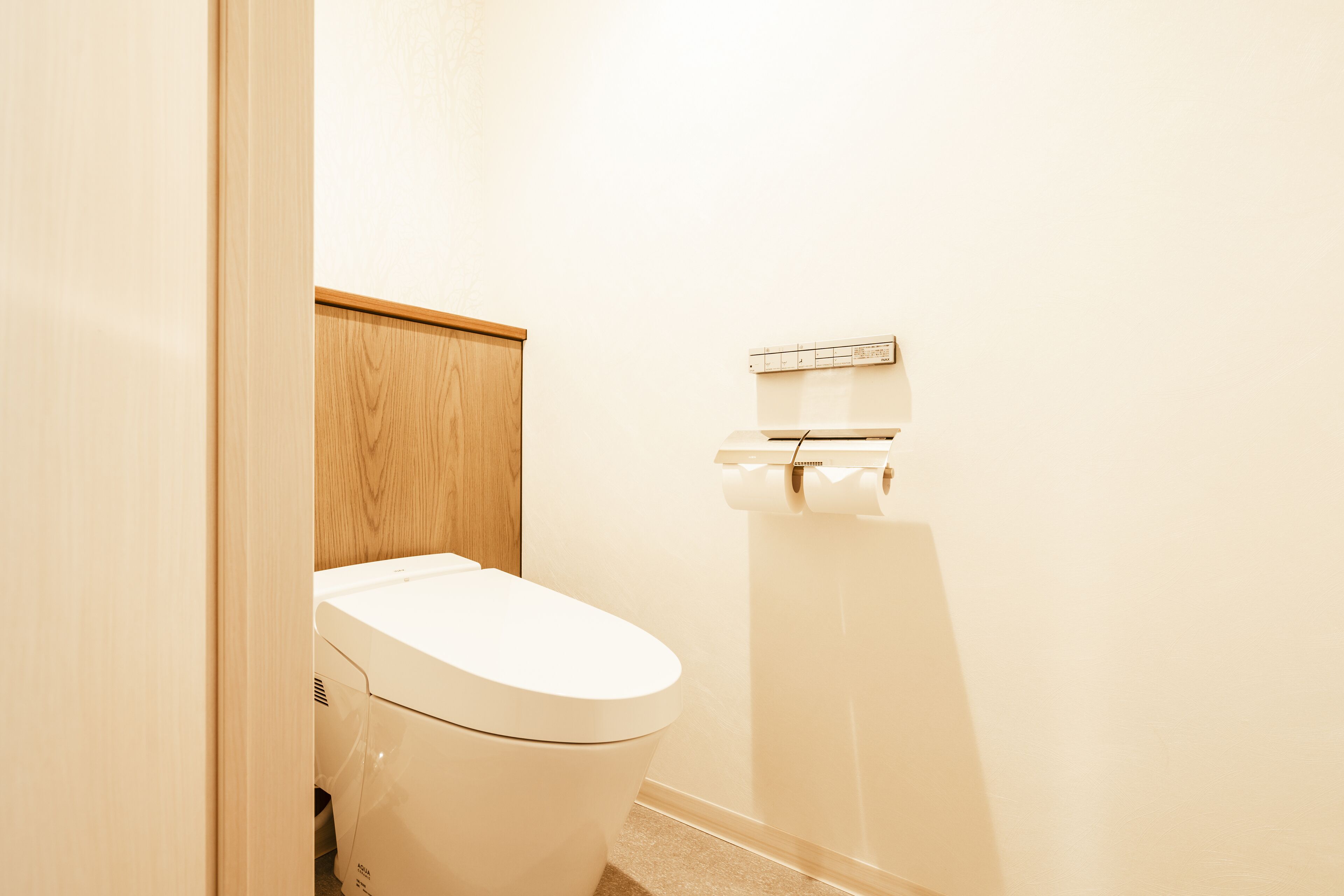 A white toilet with a wooden panel above it, a toilet paper holder on the wall, and a beige floor.