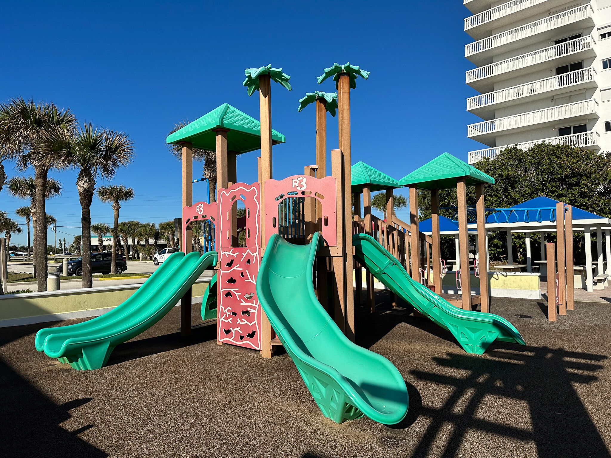 Un parque infantil con dos toboganes y una estructura para escalar, rodeado de palmeras y un edificio al fondo.