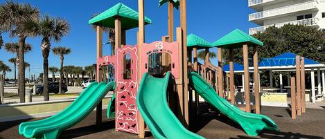 Un parque infantil con dos toboganes y una estructura para escalar, rodeado de palmeras y un edificio al fondo.
